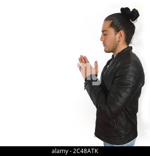 Young hispanic man with gathered hair done bow dressed in black shirt and black leather jacket, with his hands clasped in prayer position, seen in pro Stock Photo