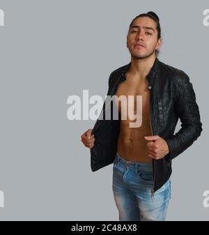 Young hispanic man with gathered hair done bow in black leather jacket without shirt, with his hands stretching his jacket, looking towards the camera Stock Photo