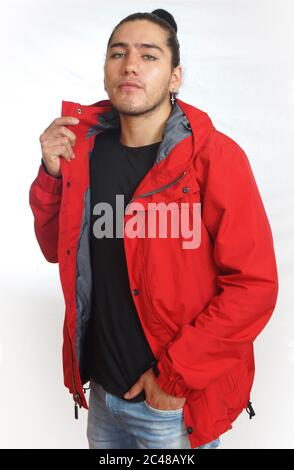 Young hispanic man with gathered hair done in bun wearing black t-shirt and red jacket, with one hand on his pants pocket, looking towards the camera Stock Photo