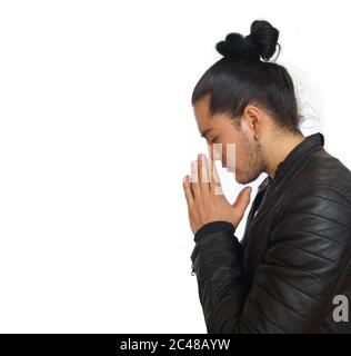 Young hispanic man with gathered hair done bow wearing black t-shirt and black leather jacket, with his hands clasped in prayer position with crouched Stock Photo