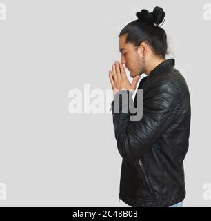 Young hispanic man with gathered hair done bow wearing black t-shirt and black leather jacket, with his hands clasped in prayer position with crouched Stock Photo