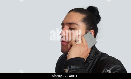 Young hispanic man with gathered hair done bow in black dress and black leather jacket, using mobile phone, seen in profile on white background Stock Photo