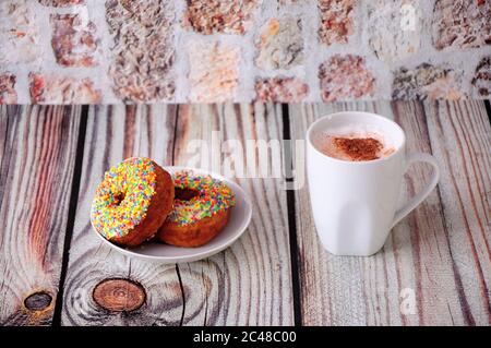 Two fresh donuts in colorful sprinkles and a cup of coffee stand on a wooden table against a stone wall. Close-up. Stock Photo