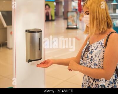 Portrait of beautiful woman wearing protective face mask uses automatic hand sanitizer dispenser at the entrance of shopping center during covid-19 ou Stock Photo