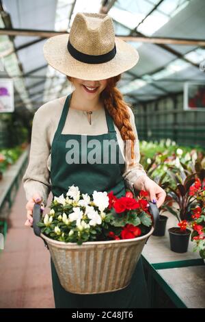 Smiling florist in apron and hat standing and holding flowers in metal basket while working in greenhouse Stock Photo
