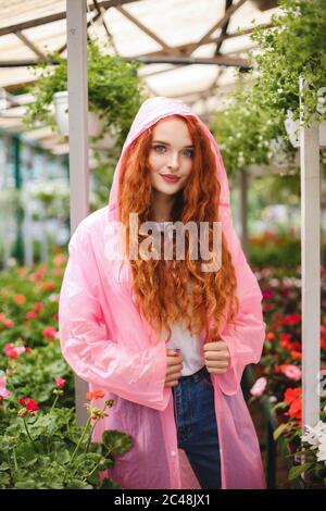Beautiful lady with redhead curly hair standing in pink raincoat and dreamily looking in camera while spending time in greenhouse Stock Photo