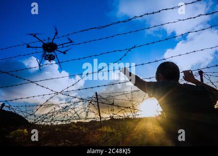 Man looking through barbed wire fence at sunrise with drone overhead. Brexit, immigration, asylum, people smuggling, border control... concept. Stock Photo