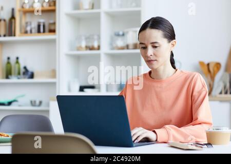 Young woman using laptop computer at her work while sitting at the table in the kitchen Stock Photo