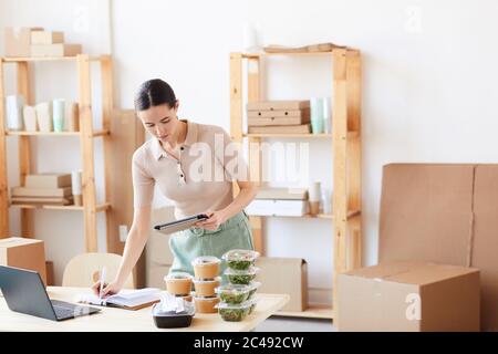 Young woman making notes in note pad and using digital tablet she making a delivery at office Stock Photo