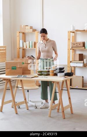 Young woman using digital tablet and packing food in packages while working at office in delivery service Stock Photo