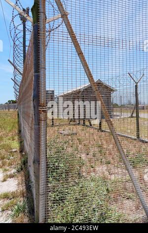 Robben Island, South Africa - 24 November 2019: Wire mesh and razor wire fence surrounding one of the prison compounds. Some buildings are on the outs Stock Photo