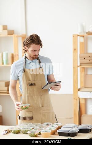 Young man in apron using tablet pc and packing boxes with fresh vegetables for delivery Stock Photo