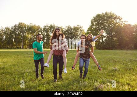 Millennial friends having fun outdoors on summer weekend. Two young guys giving their female friends piggyback ride Stock Photo
