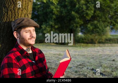 Closeup shot of a young male in a red suit reading a book sitting and leaning on a tree Stock Photo