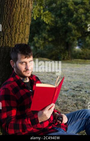 Closeup shot of a young male in a red suit reading a book sitting and leaning on a tree Stock Photo