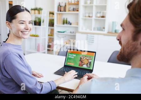 Young woman smiling to man and making food delivery on laptop while they sitting at the table in the kitchen Stock Photo