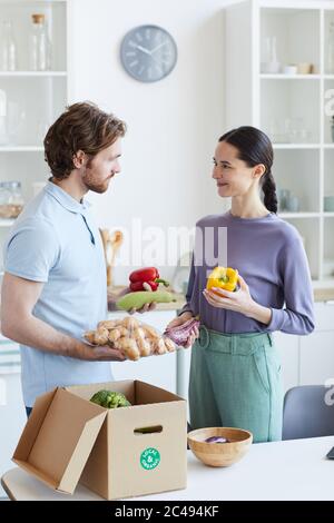 Young couple taking fresh vegetables out of the box and talking to each other while standing in the kitchen Stock Photo