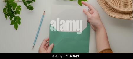 Overhead shot of female hands open mock-up greeting card with turquoise  envelope on table with pencil and decorations Stock Photo