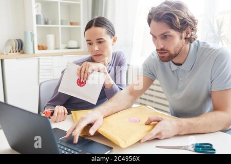 Young couple sitting at the table with envelopes and parcels and checking their email on laptop at home Stock Photo