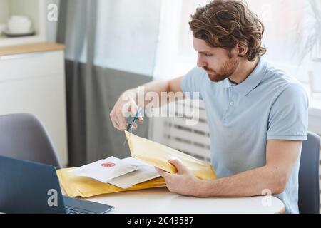 Young man sitting at the table in front of laptop and opening the paper envelopes at home Stock Photo