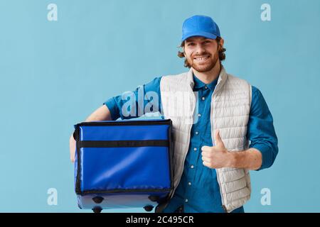 Portrait of young man working in delivery service he smiling at camera and showing thumb up against the blue background Stock Photo