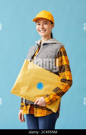 Portrait of young postwoman in uniform holding big parcel and smiling at camera isolated on blue background Stock Photo