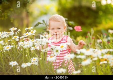 Baby girl smiles in summer dress walking outside among daisies in garden Stock Photo