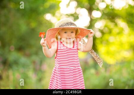 Cute little girl in a red dress and hat walks in summer barefoot on the green grass, space for text Stock Photo