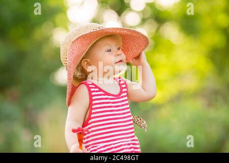 Cute little girl in a red dress and hat walks in summer barefoot on the green grass, space for text Stock Photo