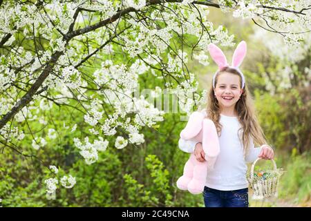 Happy child walking in garden with blossoming trees. Girl smiling in bunny ears, rabbit and basket. Childhood, youth and growth. Easter and spring concept. Holidays celebration with presents and toys. Stock Photo