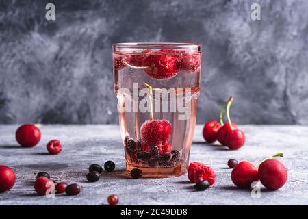 Fresh cold sparkling water with cherry, raspberry and currant berries in red faceted glass on stone concrete background, summer diet beverage, angle v Stock Photo