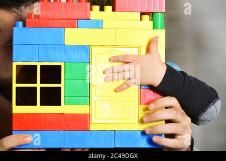 Dad and kid hide behind building wall made of plastic blocks. Guys play together on defocused background. Family game and childhood concept. Childs and male hands hold colorful toy bricks construction Stock Photo