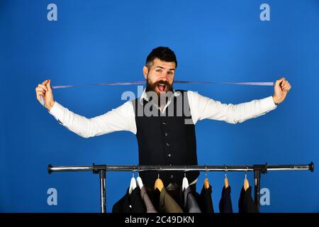 Man with beard by clothes rack. Fashion and male style concept. Tailor with happy face holds measuring tape near jackets on blue background. Designer takes measures near clothes hangers with suits Stock Photo