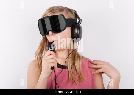 Cute girl wearing 3D VR glasses and headphones and holding a microphone sings a song in a virtual live concert Stock Photo