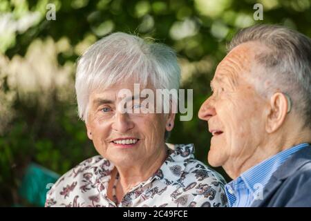 Head-and-shoulder portrait of an elderly pair of reindeer in front of green blurred leavesin summer. Woman with white hair looks at camera. Stock Photo