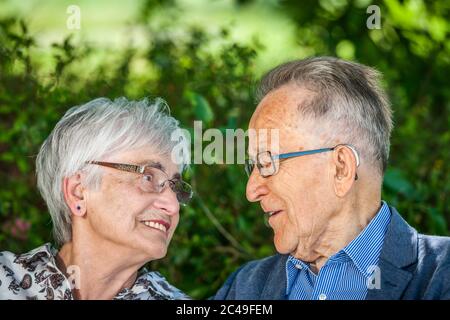 Head-and-shoulders portrait of an elderly pensioner couple looking into each other's eyes with glasses in front of green leaves in summer. Stock Photo