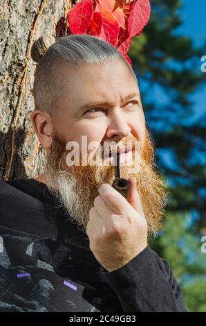 Guy smoking tobacco pipe. Hipster with red beard and mustache standing near tree trunk in the park. Bad habits concept. Vertical orientation Stock Photo