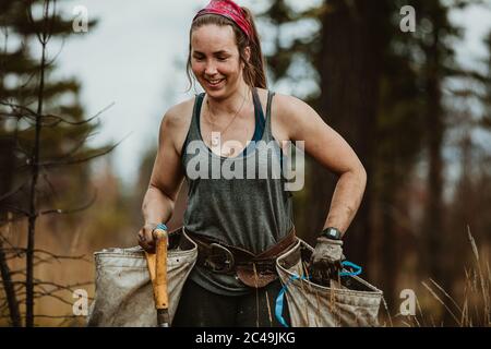 Woman planting trees in forest. Female tree planter carrying bags full of trees and a shovel. Stock Photo