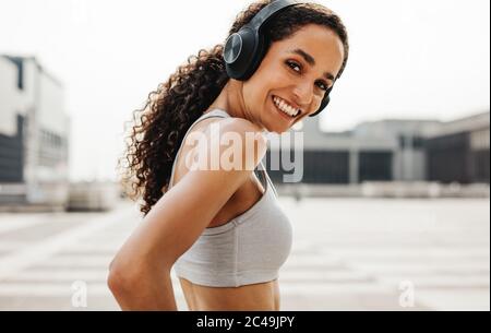 Portrait of beautiful fitness woman wearing headphones looking at camera and smiling. Woman in sportswear exercising outdoors in the city. Stock Photo