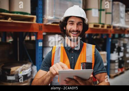 Handsome cheerful young caucasian smiling supervisor with white helmet using digital tablet for work while standing in warehouse against goods Stock Photo