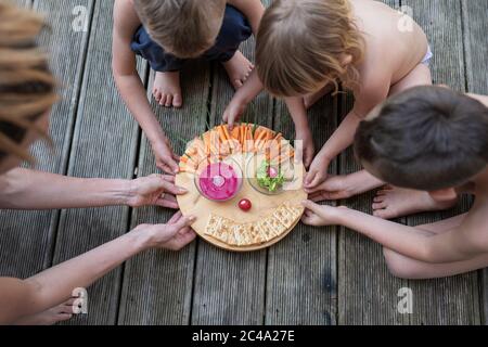 Top view of a mother and her three children holding a round cutting board with a vegan snack of raw carrots, crackers and dips on it. Stock Photo
