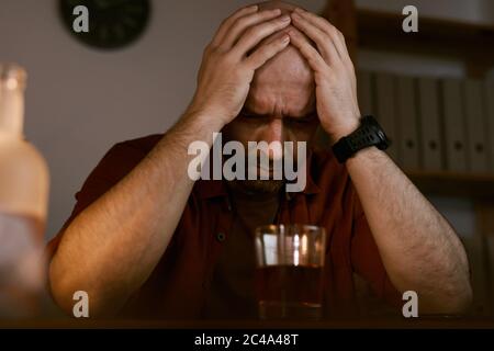 Upset mature man sitting at the table and holding his head with hands he has problems with alcohol Stock Photo