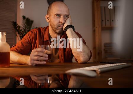 Mature man sitting at the table in front of computer and thinking about something while drinking alcohol Stock Photo