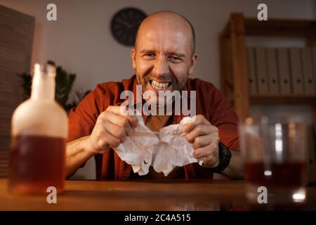 Portrait of mature angry man sitting at the table with bottle of wine and tearing the letter Stock Photo