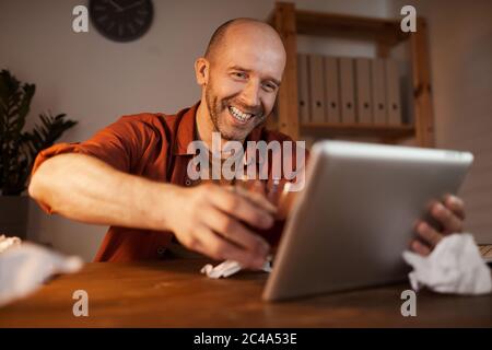 Mature man sitting at the table and using digital tablet for online conversation he talking to his friend and drinking alcohol Stock Photo