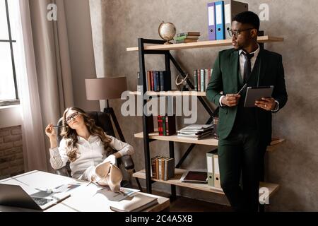 Work in a creative office. An African man and a beautiful business lady in formal clothes are working together, standing indoors Stock Photo