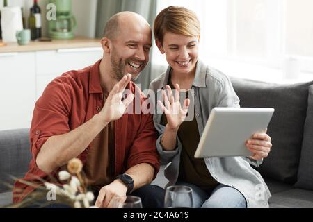 Young happy couple sitting on sofa and using digital tablet for online conversation they waving and smiling Stock Photo