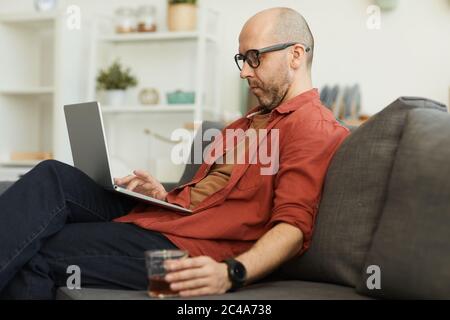 Mature man sitting on sofa using laptop computer and drinking whiskey he working at home Stock Photo