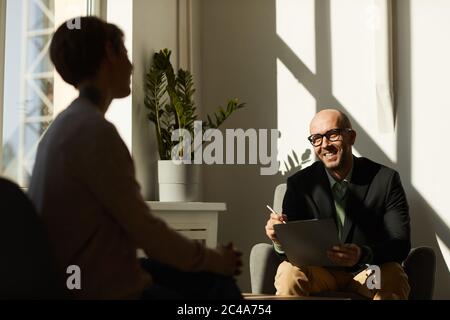 Mature man in eyeglasses sitting on sofa with laptop and smiling while talking to woman at office Stock Photo