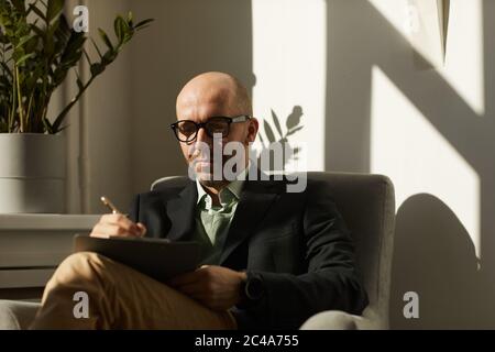 Mature serious businessman in eyeglasses sitting on armchair and making notes in document he working at office Stock Photo
